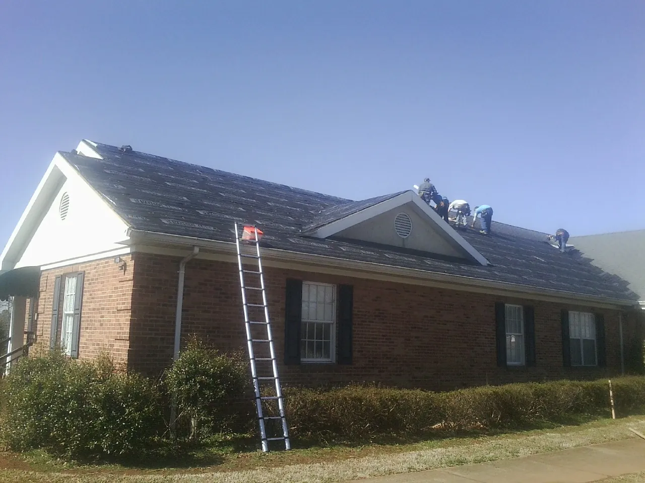 Crew tearing shingles off a brick ranch roof with a ladder set — Emergency Roof Repair work by our crew in Ashburnham