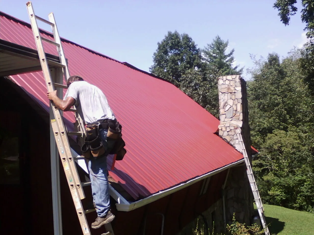 Roofer on a ladder beside a red metal roof and stone chimney — Soffit & Fascia Repair by Roofing Brothers in Ashburnham
