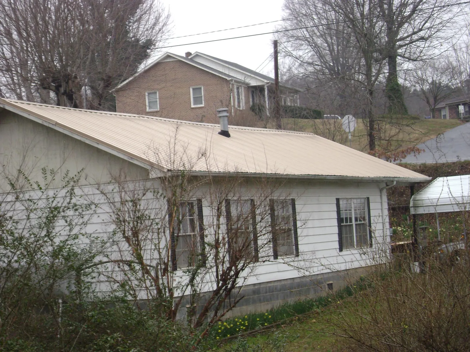 Tan metal roof on a white sided house set into a hillside — Commercial Roofing by Roofing Brothers in Ashburnham